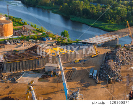 A construction site near a river in Riga, Latvia, with a partially built bridge, cranes, vehicles, scrap metal, and industrial tanks under warm sunlight. A construction site near a river in Riga, Latvia, with a partially built bridge, cranes, vehicles, scrap metal, and industrial tanks under warm sunlight. 129560874