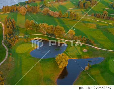 A lush green golf course at sunset featuring fairways, sand bunkers, and water hazards. A winding path and trees add depth, with water in the background. A lush green golf course at sunset featuring fairways, sand bunkers, and water hazards. A winding path and trees add depth, with water in the background. 129560875