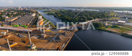 A river winds through greenery with a bridge under construction, industrial buildings, cranes, and Riga's urban skyline in the distance at golden hour. A river winds through greenery with a bridge under construction, industrial buildings, cranes, and Riga's urban skyline in the distance at golden hour. 129560886