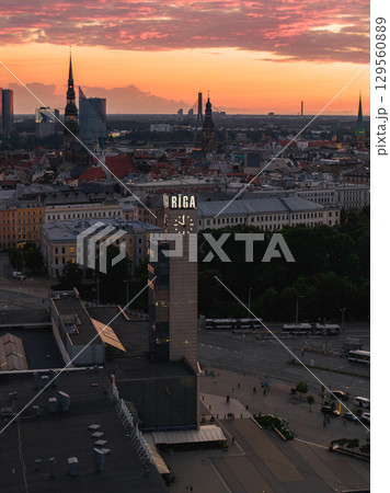 Riga clock tower in the foreground with St. Peter's Church spire and old town skyline at sunset. Warm hues highlight urban activity and architecture. 129560889