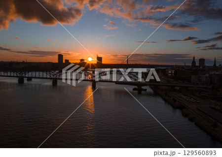 The sun sets over Riga, Latvia, with the Railway Bridge spanning the Daugava River. The Riga TV Tower and old town skyline are visible in the distance. 129560893