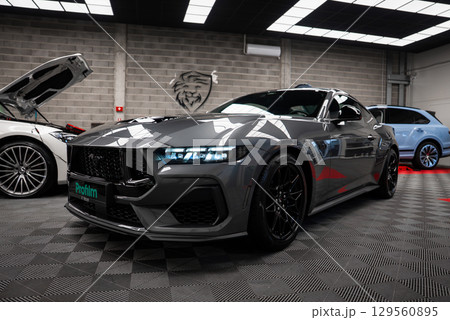A gray Ford Mustang GT 5.0 with black alloy wheels in a bright indoor showroom. A Mercedes AMG G Class and Bentley are partially visible in the background. 129560895