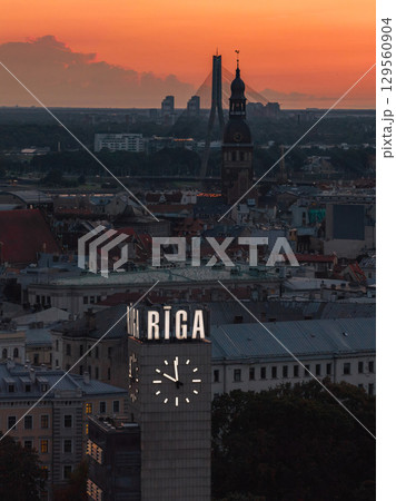 Riga clock tower with illuminated sign and clock face at sunset, St. Peter's Church spire, and Vansu Bridge spanning the Daugava River in Latvia. 129560904