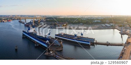 A shipyard in Riga, Latvia, during sunset. Two large ships in dry docks are surrounded by cranes, industrial structures, and the Daugava River. 129560907