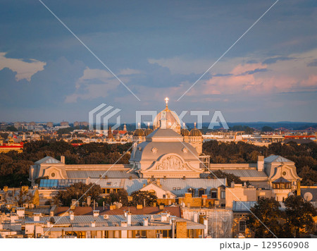 Golden sunset light illuminates the Nativity of Christ Cathedral in Riga, Latvia, with its domes and cross, surrounded by rooftops and a tree lined background. 129560908