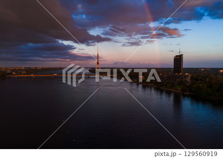 View of Riga, Latvia, featuring the Riga TV Tower, a faint rainbow, the Daugava River, a bridge, modern buildings, and dramatic sunset clouds. 129560919