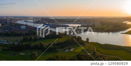 Serene sunset over Riga, Latvia, featuring a river, a bridge, a shipyard with docked vessels, lush greenery, and a golf course with visible fairways. Serene sunset over Riga, Latvia, featuring a river, a bridge, a shipyard with docked vessels, lush greenery, and a golf course with visible fairways. 129560929