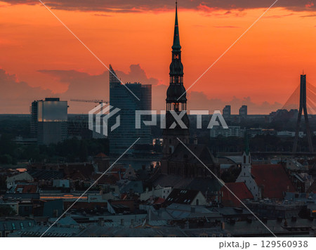 Silhouette of St. Peter's Church spire at sunset in Riga, Latvia, with old town rooftops, Vansu Bridge, and modern buildings in the background. 129560938