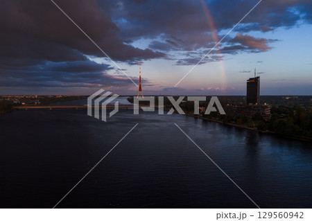 Serene sunset over the Daugava River in Riga, Latvia, featuring the Riga TV Tower, a bridge, modern buildings, greenery, and a partial rainbow. Serene sunset over the Daugava River in Riga, Latvia, featuring the Riga TV Tower, a bridge, modern buildings, greenery, and a partial rainbow. 129560942