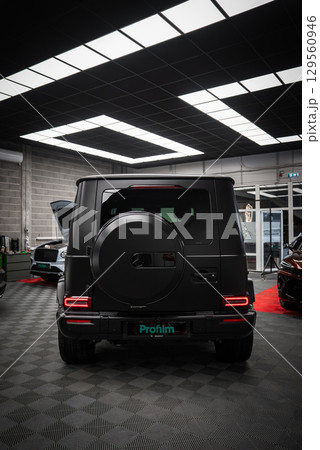 Matte black Mercedes AMG G Class SUV parked indoors on a checkered floor. Spare tire cover, taillights, and nearby vehicles visible in a modern showroom. 129560946