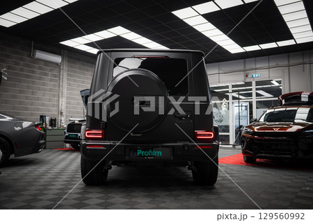 Matte black Mercedes AMG G Class SUV with spare tire cover and taillights in a well lit showroom. Ford Mustang GT 5.0 and red car partially visible. Matte black Mercedes AMG G Class SUV with spare tire cover and taillights in a well lit showroom. Ford Mustang GT 5.0 and red car partially visible. 129560992