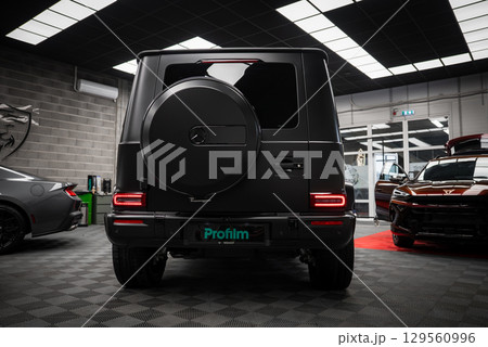 Black Mercedes AMG G Class SUV with spare tire cover, parked indoors on checkered floor. Gray Ford Mustang and red Cupra partially visible nearby. 129560996