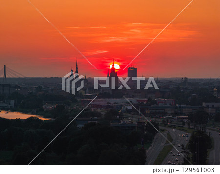 Sunset over Riga, Latvia, with the Latvian Academy of Sciences spire silhouetted, Vansu Bridge over the Daugava River, and lush greenery in the foreground. 129561003