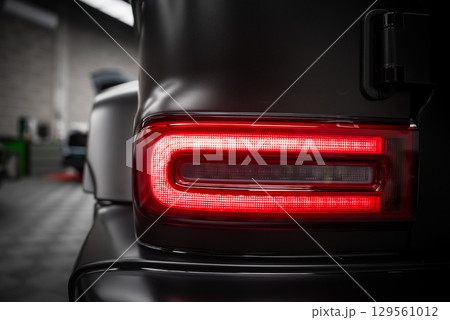 Close up view of the Mercedes AMG G Class rear taillight with rectangular LED design, matte black exterior, and blurred indoor showroom background. 129561012