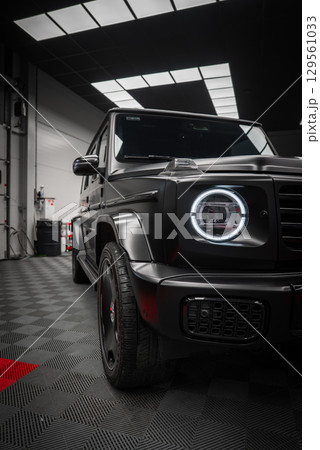 Black Mercedes AMG G Class SUV with matte finish, circular LED headlight, and bold grille in a modern showroom with patterned black floor. Black Mercedes AMG G Class SUV with matte finish, circular LED headlight, and bold grille in a modern showroom with patterned black floor. 129561033