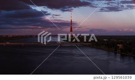Riga TV Tower stands tall during sunset with purple and pink skies. The Daugava River flows below, crossed by a bridge with light vehicle traffic. 129561039