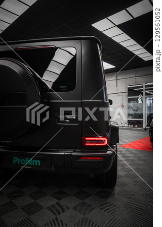 Matte black Mercedes AMG G Class parked indoors on a checkered floor with red carpet, showcasing spare tire cover, LED taillights, and Profilm logo. 129561052