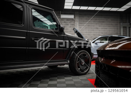 A black Mercedes AMG G Class and part of a Ford Mustang GT 5.0 in a modern showroom with gray brick walls, checkered floor, and bright lighting. 129561054