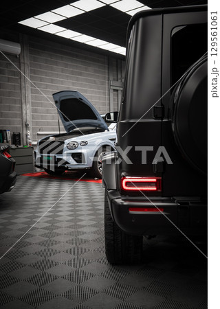 A matte black Mercedes AMG G Class with visible taillights and spare tire cover in a modern showroom. A white Bentley with hood open is in the background. 129561061
