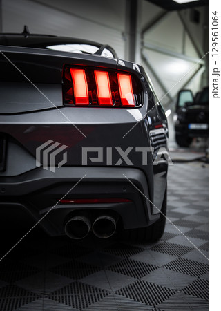 Close up of Ford Mustang GT 5.0 rear end with illuminated taillights and dual exhaust pipes. Parked indoors on a checkered floor with a Mercedes AMG G Class visible. 129561064