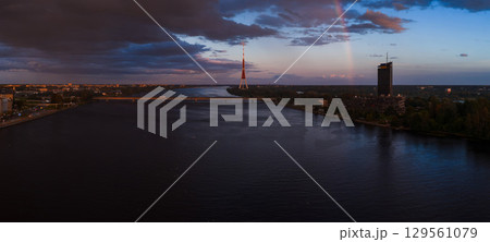 View of the Daugava River in Riga, Latvia, featuring the Riga TV Tower, a faint rainbow, a bridge, modern architecture, and dramatic sunset clouds. 129561079