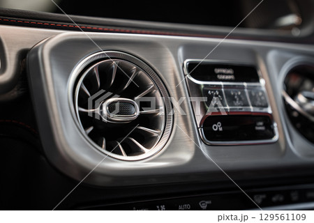 Close up view of a Mercedes AMG G Class interior, highlighting a turbine style air vent, red stitched dashboard trim, and sleek climate controls. 129561109