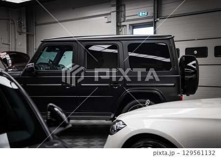 A sleek black Mercedes AMG G Class SUV with a matte finish parked indoors. Features include its boxy design, rear spare tire, and polished surroundings. 129561132