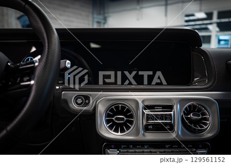 Close up of Mercedes AMG G Class dashboard with circular air vents, central display, carbon fiber accents, and leather stitching in a showroom. 129561152