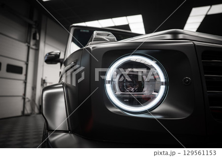 Close up of a Mercedes AMG G Class headlight with circular LED design, matte black finish, and angular body lines in a tiled indoor showroom. Close up of a Mercedes AMG G Class headlight with circular LED design, matte black finish, and angular body lines in a tiled indoor showroom. 129561153