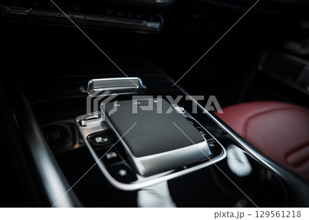 Detailed view of a Mercedes AMG G Class center console featuring a touchpad controller, metallic buttons, and a red leather seat in the background. 129561218