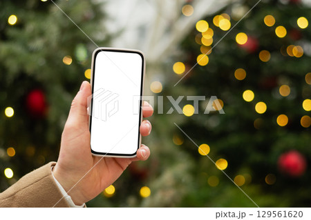 Man hand with smartphone with white blank screen against Christmas tree light garland and fairy lights. Festive Xmas Background. Mock up. Close up. 129561620