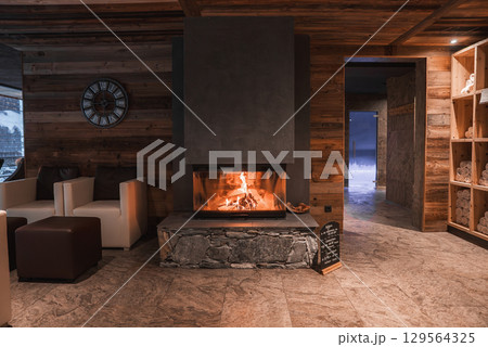 Warm room with wooden walls, modern fireplace, armchairs, and spa shelves. A doorway reveals stone walls and a snowy outdoor view. Warm room with wooden walls, modern fireplace, armchairs, and spa shelves. A doorway reveals stone walls and a snowy outdoor view. 129564325