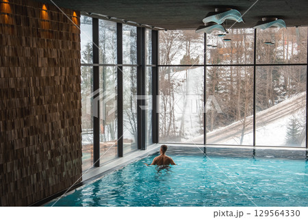 A person swims in an indoor pool with large windows revealing a snowy forest. The space features wooden walls and modern light fixtures, set in the Italian Alps. A person swims in an indoor pool with large windows revealing a snowy forest. The space features wooden walls and modern light fixtures, set in the Italian Alps. 129564330