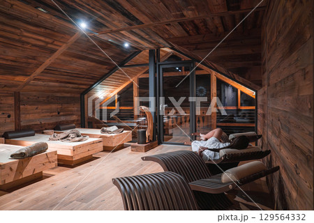 Cozy relaxation room in a luxury hotel in the Italian Alps, featuring sloped wooden ceilings, modern lounge chairs, and large glass doors to a balcony. Cozy relaxation room in a luxury hotel in the Italian Alps, featuring sloped wooden ceilings, modern lounge chairs, and large glass doors to a balcony. 129564332