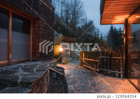 Cozy outdoor space of a luxury hotel in the Italian Alps, featuring a stone pathway, warm lighting, rustic wooden fence, and hilly backdrop with trees. Cozy outdoor space of a luxury hotel in the Italian Alps, featuring a stone pathway, warm lighting, rustic wooden fence, and hilly backdrop with trees. 129564354