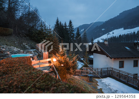 Chalet style building with sloped roof, wooden balcony, and illuminated hot tub. Snow patches, evergreen trees, and forested hills in the background. 129564358