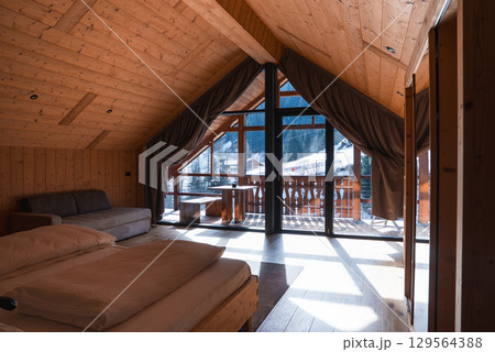 Wooden interior of a hotel room with sloped ceiling, large windows, and balcony overlooking snow covered rooftops and forested mountains. 129564388