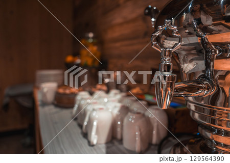 Cozy indoor scene with a metal beverage dispenser, white ceramic cups, pastries, orange juice, wooden walls, and warm lighting in the Italian Alps. Cozy indoor scene with a metal beverage dispenser, white ceramic cups, pastries, orange juice, wooden walls, and warm lighting in the Italian Alps. 129564390
