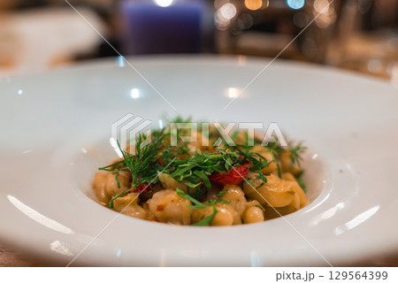 Close up of pasta garnished with dill, parsley, and red chili in a white bowl. Warm lighting and a purple candle create a cozy atmosphere. Close up of pasta garnished with dill, parsley, and red chili in a white bowl. Warm lighting and a purple candle create a cozy atmosphere. 129564399