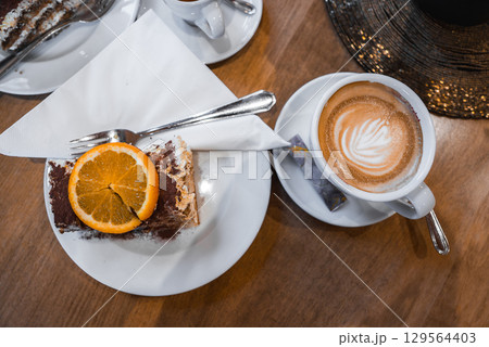 A slice of tiramisu with orange garnish on a white plate, paired with a cappuccino featuring latte art, set on a wooden table in a luxury setting. A slice of tiramisu with orange garnish on a white plate, paired with a cappuccino featuring latte art, set on a wooden table in a luxury setting. 129564403