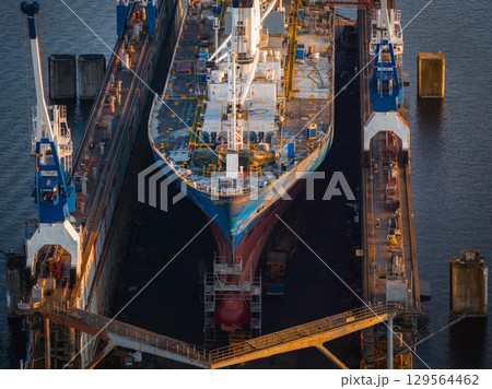 A large ship in a dry dock at Riga shipyard, surrounded by blue and white cranes. The hull features vibrant colors, with scaffolding and equipment visible. A large ship in a dry dock at Riga shipyard, surrounded by blue and white cranes. The hull features vibrant colors, with scaffolding and equipment visible. 129564462