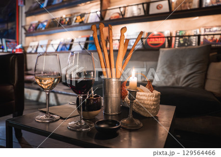 Indoor lounge with a table set with wine glasses, breadsticks, and a candle. Shelves of vinyl records and soft lighting create a retro ambiance. Indoor lounge with a table set with wine glasses, breadsticks, and a candle. Shelves of vinyl records and soft lighting create a retro ambiance. 129564466