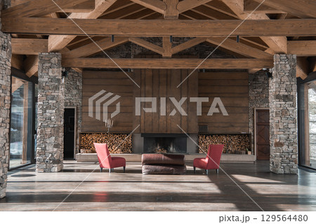 Grand interior of an alpine hotel featuring a central fireplace, exposed wooden beams, stone walls, red armchairs, and large windows with mountain views. Grand interior of an alpine hotel featuring a central fireplace, exposed wooden beams, stone walls, red armchairs, and large windows with mountain views. 129564480