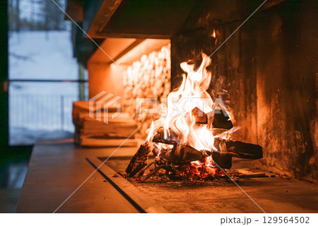 Cozy indoor fireplace with stacked firewood, warm lighting, and a large window showcasing a snowy winter scene in the Italian Alps. 129564502