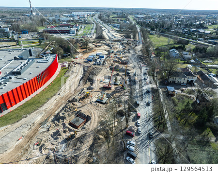 Large construction site in Riga, Latvia, with cranes, excavators, and materials. A red curved building, busy road, and residential areas are visible. 129564513