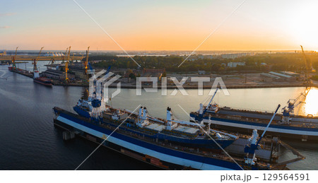 Vibrant sunset over a Riga shipyard with blue and white ships docked, cranes along the waterfront, and urban structures in the background. 129564591