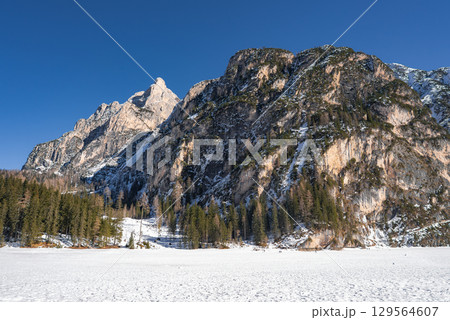Winter scene at Braies Lake, Italy, featuring a frozen lake, rocky snow dusted cliffs, evergreen trees, and a sharp mountain peak under a clear sky. 129564607