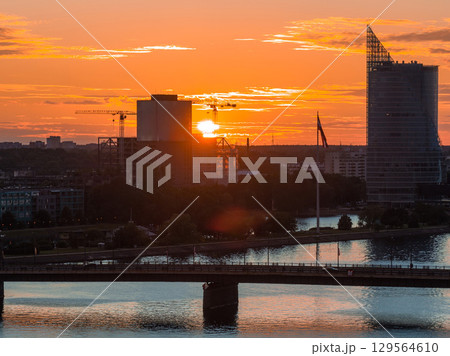 Warm sunset over Riga, Latvia, with the Daugava River reflecting orange hues, a bridge in the foreground, and a triangular skyscraper on the right. 129564610