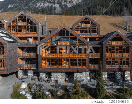 Charming Alpine hotel with steep roofs and wooden balconies, surrounded by forested slopes, rocky peaks, and patches of snow in the Dolomites, Italy. 129564626