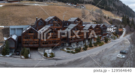 Cluster of wooden Alpine style buildings with sloped roofs and balconies, set in a mountainous area with a paved road, parking, and hillside trees. Cluster of wooden Alpine style buildings with sloped roofs and balconies, set in a mountainous area with a paved road, parking, and hillside trees. 129564640